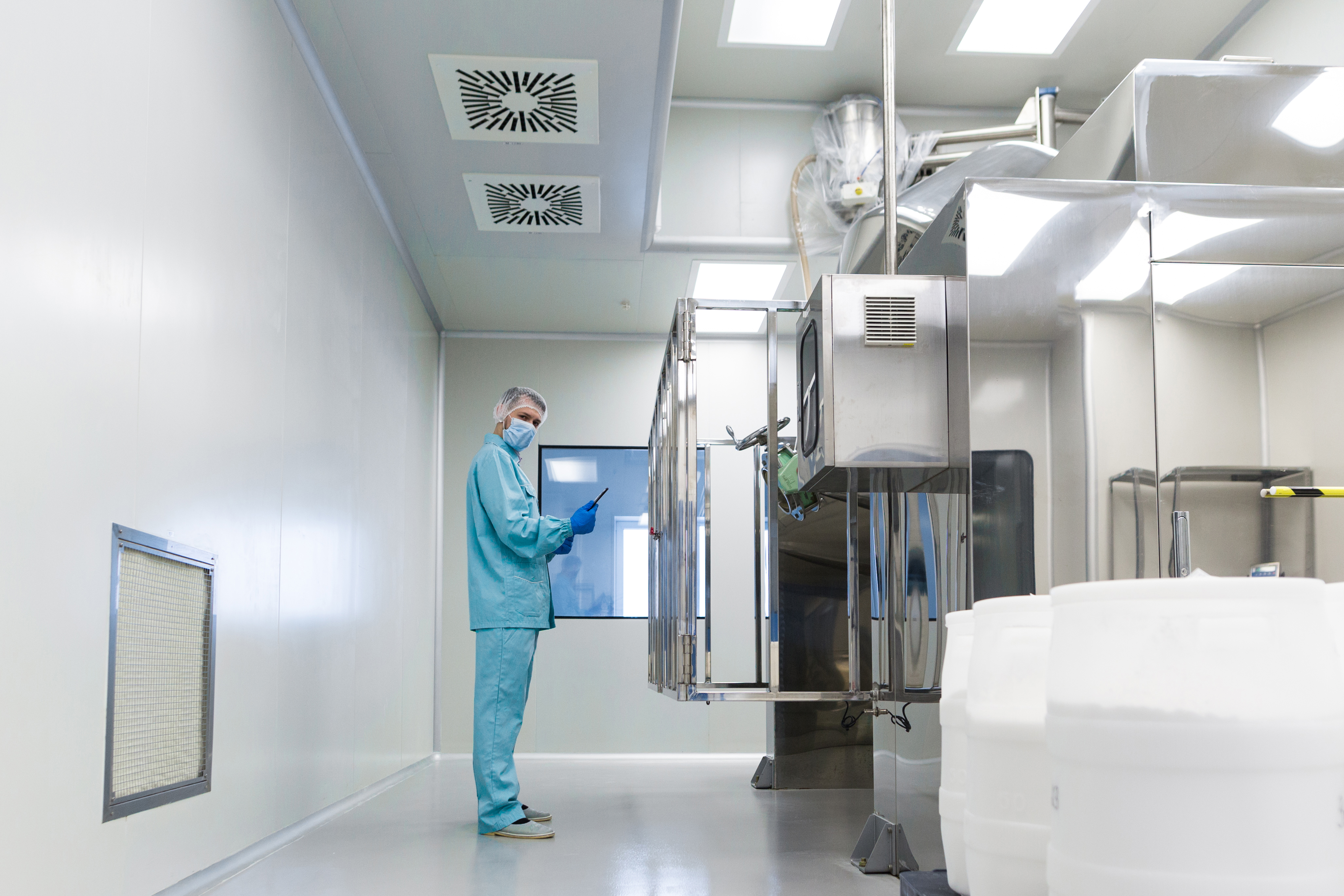 caucasian scientist in blue lab uniform make notes about big steel machine with control panel, white barrels on front, clean room, look at camera